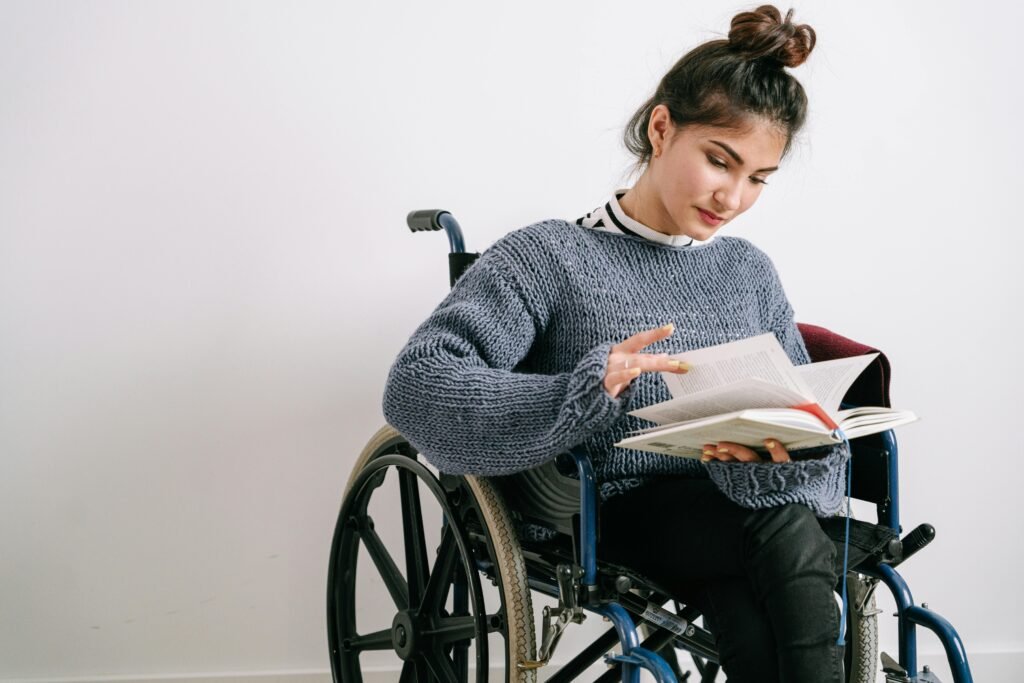 Young woman in a wheelchair reading a book indoors with a neutral background.