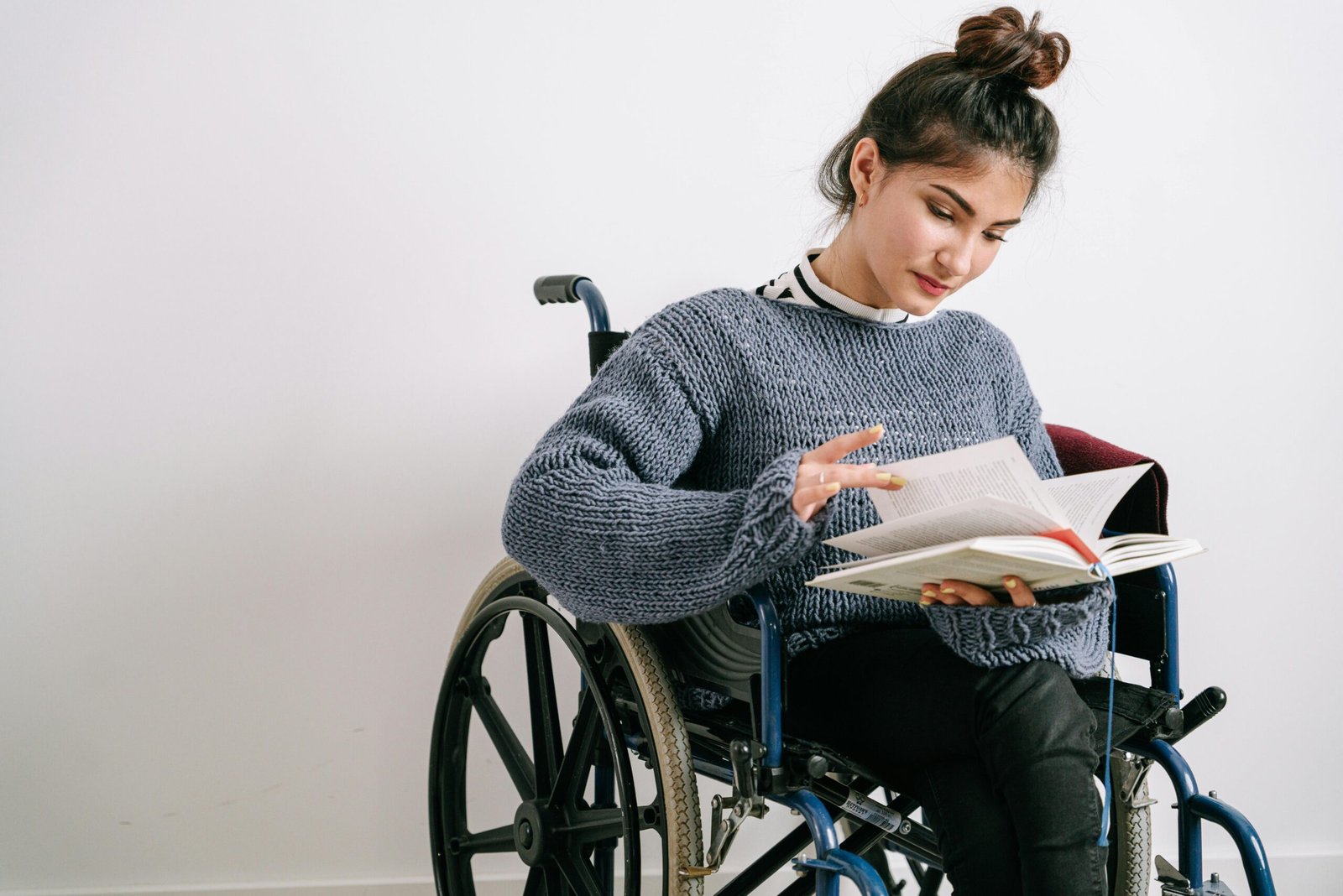 Young woman in a wheelchair reading a book indoors with a neutral background.